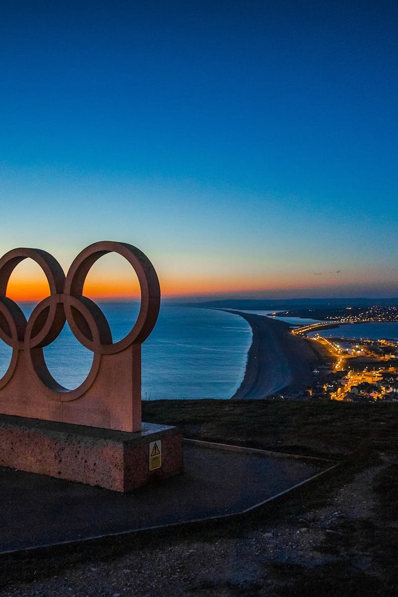 Scenic sunset view of the Olympic rings monument by the sea with city lights in the distance.
